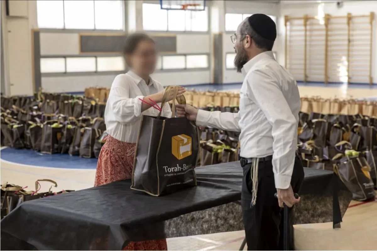 Un bénévole de Torah-Box remet un colis de Chabbath en main propre à une bénéficiaire, dans le gymnase où sont préparés les colis